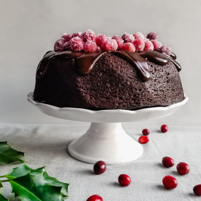 Vegan Cranberry Chocolate Fudge Cake with Ganache and Sugared Cranberries served on a white platter with holly and cranberries on the table