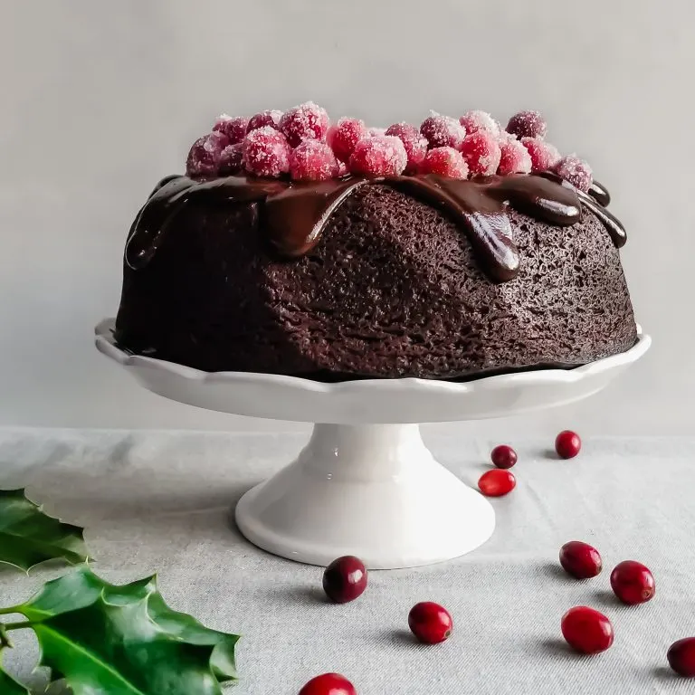 Vegan Cranberry Chocolate Fudge Cake with Ganache and Sugared Cranberries served on a white platter with holly and cranberries on the table