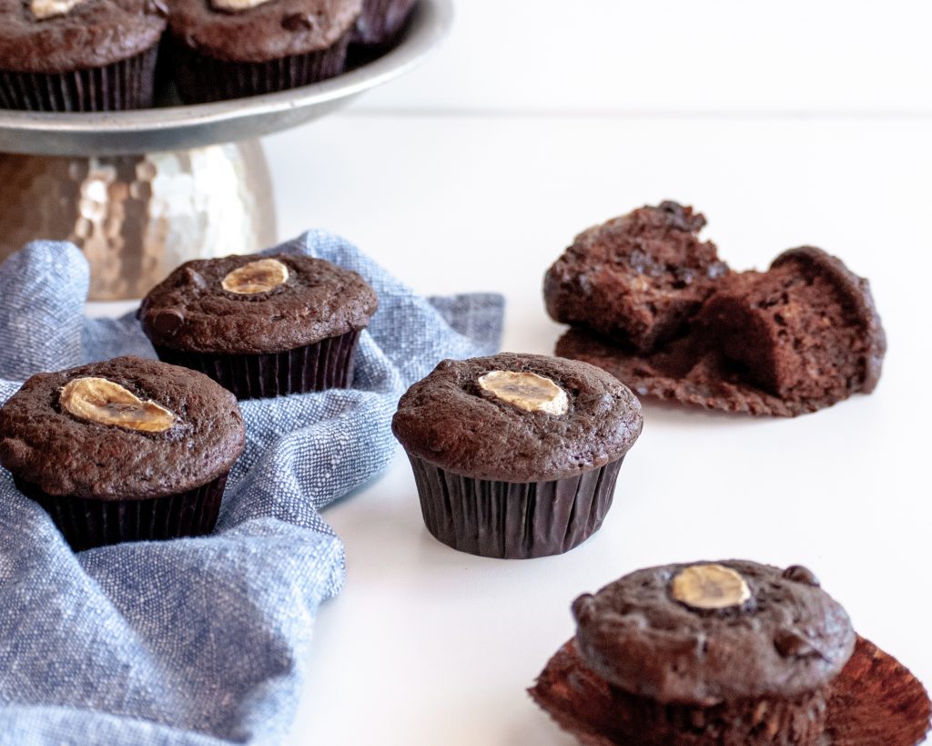 Batch of chocolate banana muffins on table, some on a platter, some with wrappers oof