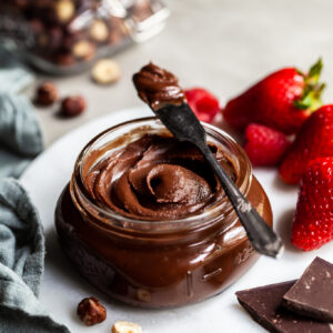 Jar filled with hazelnut chocolate spread beside fresh berries and a jar of whole hazelnuts in the background