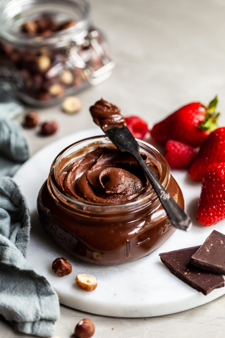 Jar filled with hazelnut chocolate spread beside fresh berries and a jar of whole hazelnuts in the background