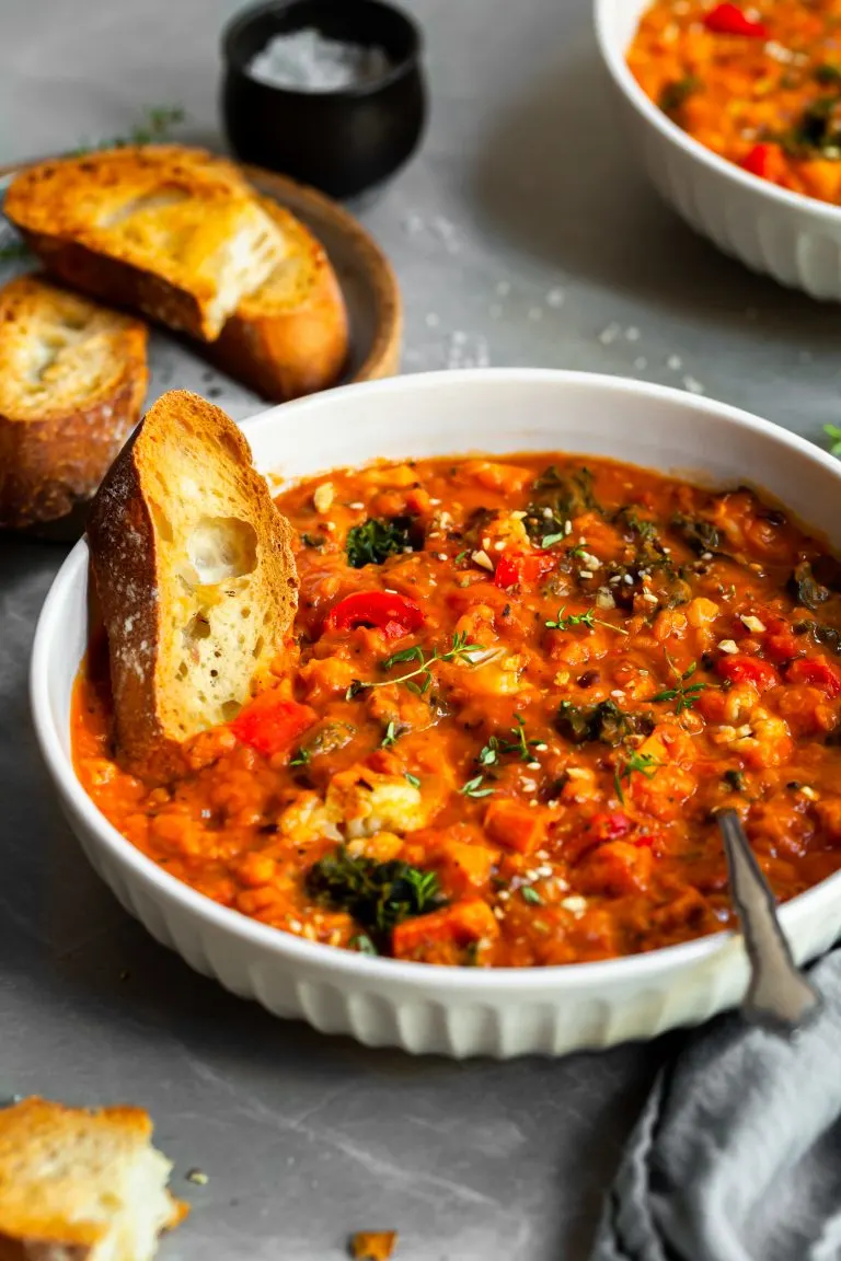 White bowl of roasted vegetable stew with red lentils and tomatoes, served with a thick slice of toast