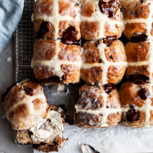 Flatlay of hot cross buns on a cooling rack, with one having been cut in half and smeared with vegan butter