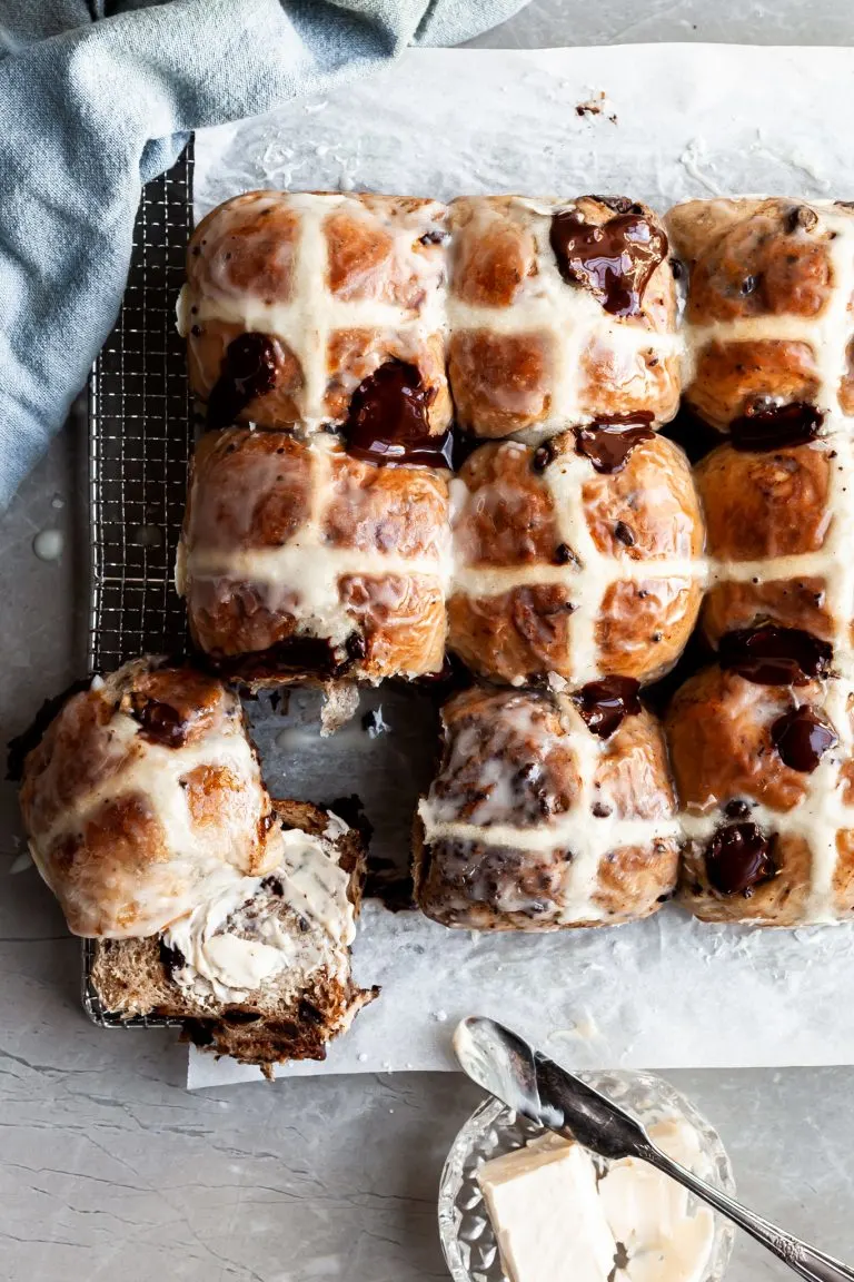 Flatlay of hot cross buns on a cooling rack, with one having been cut in half and smeared with vegan butter