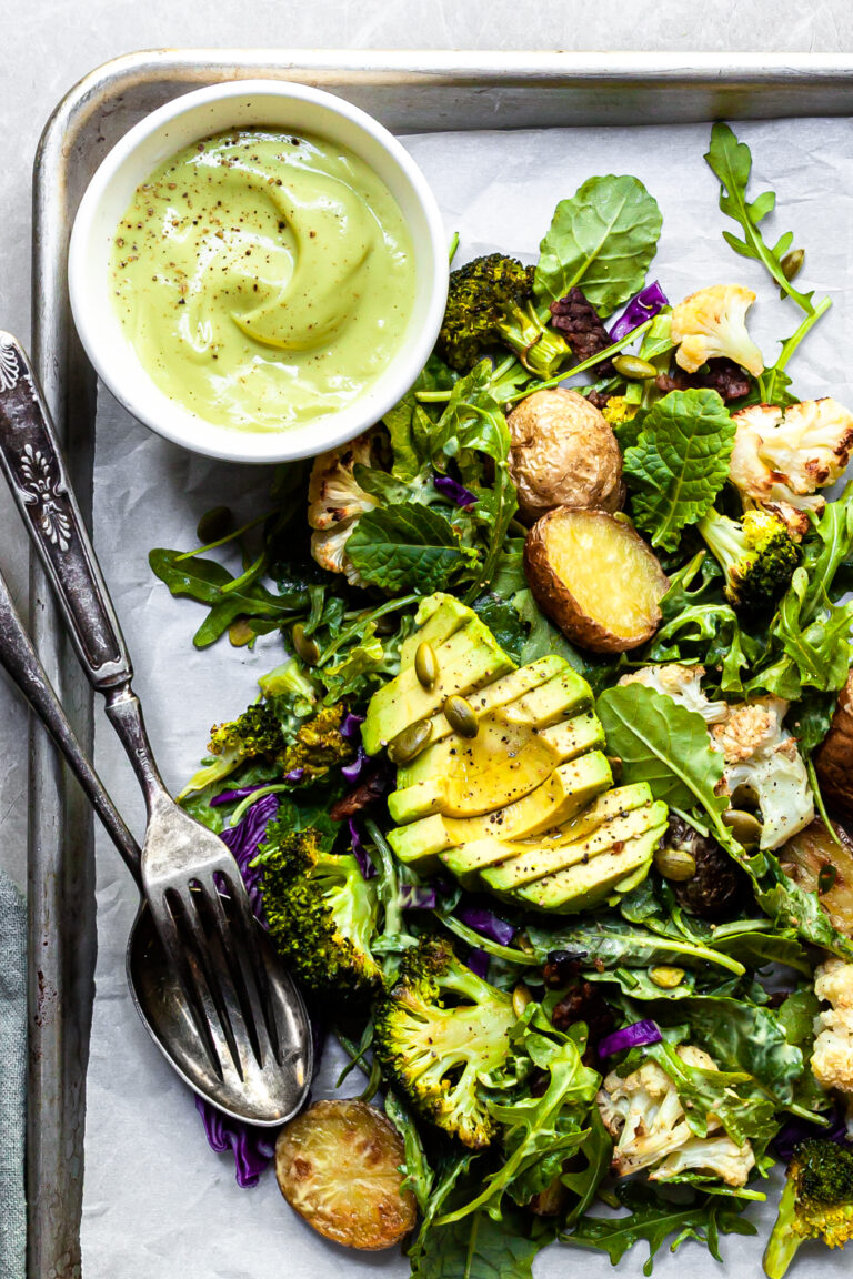 A serving of kale and roasted vegetable salad on an aluminium tray served with a small white bowl filled with creamy light green avocado Caesar dressing