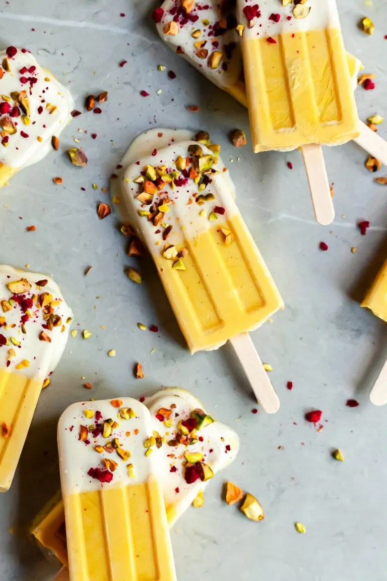 Mango Lassi Ice Pops on marble surface dipped in yogurt and sprinkled with chopped pistacios and freeze dried raspberries