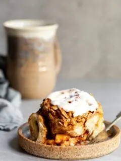 A glazed cinnamon roll on a plate with a cup of coccee in the background