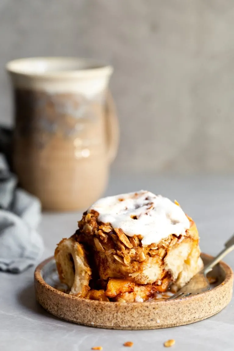 A glazed cinnamon roll on a plate with a cup of coccee in the background