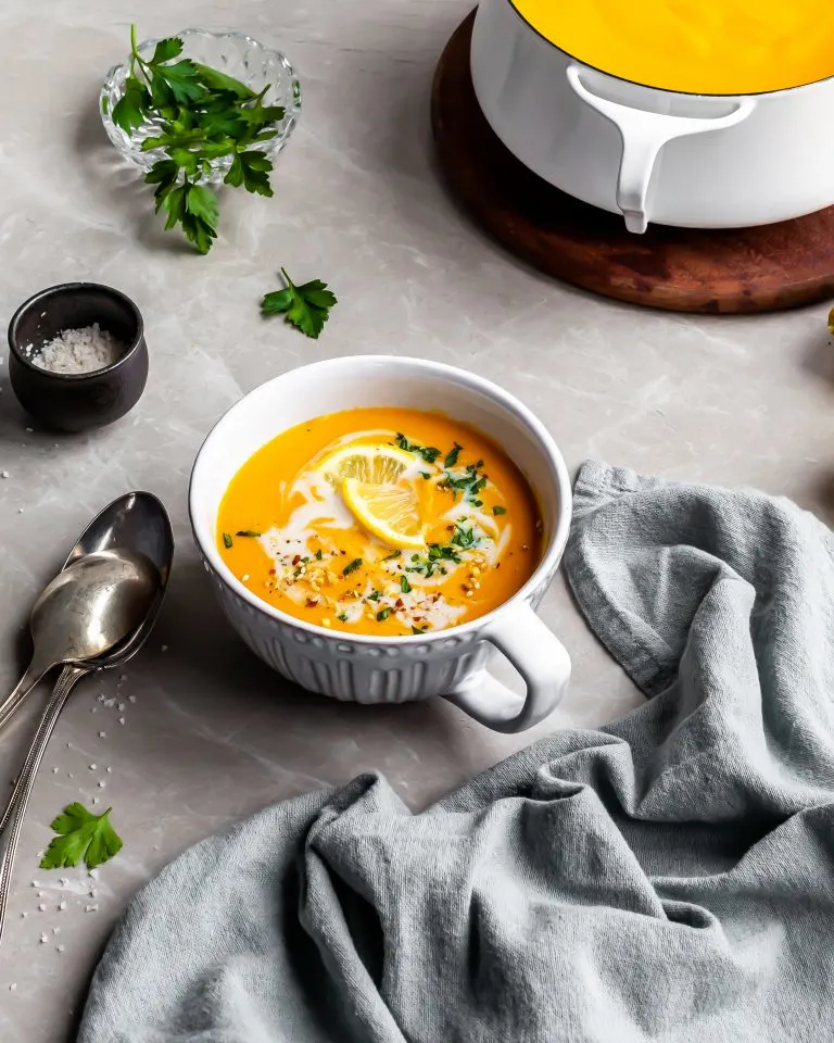 Creamy Curried Carrot, Ginger and Red Lentil Soup served in a large cup with ingredients in background