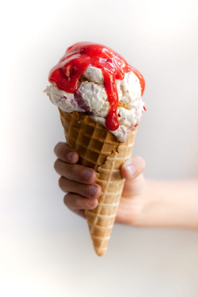 Child's hand holding a waffle cone filled with Vegan Strawberry Rhubarb Crumble Ice Cream Scoops topped with Strawberry Rhubarb Sauce