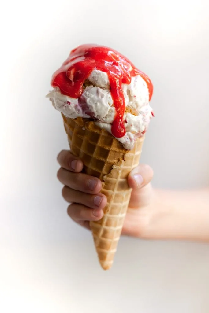 Child's hand holding a waffle cone filled with Vegan Strawberry Rhubarb Crumble Ice Cream Scoops topped with Strawberry Rhubarb Sauce