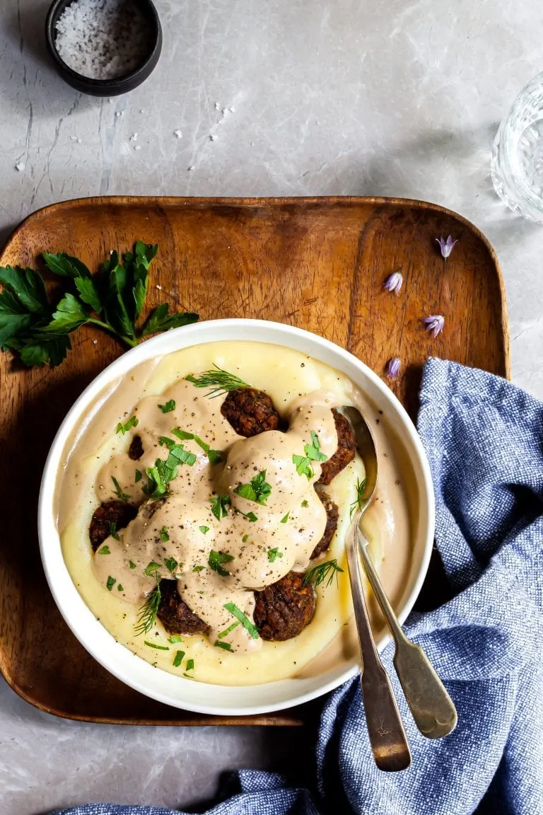 Vegan Swedish Meatballs in a bowl over mashed potatoes, covered in gravy alongside a blue napkin