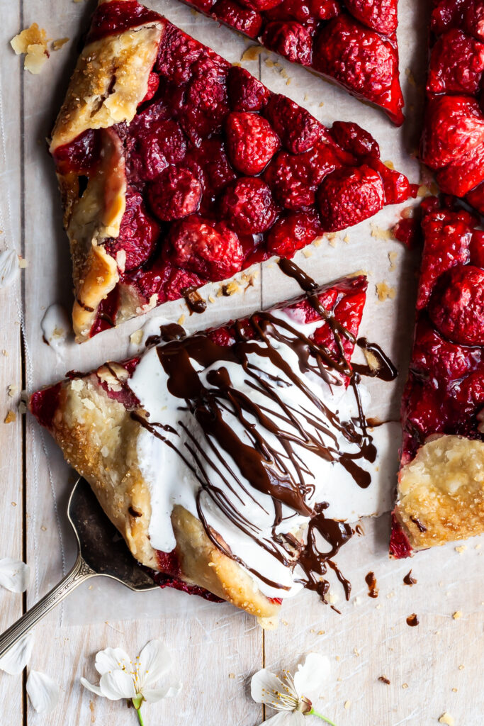 A sliced strawberry galette with one slice being served via a silver spatula. The slice is served with a scoop of ice cream on top, drizzled with chocolate hazelnut spread.