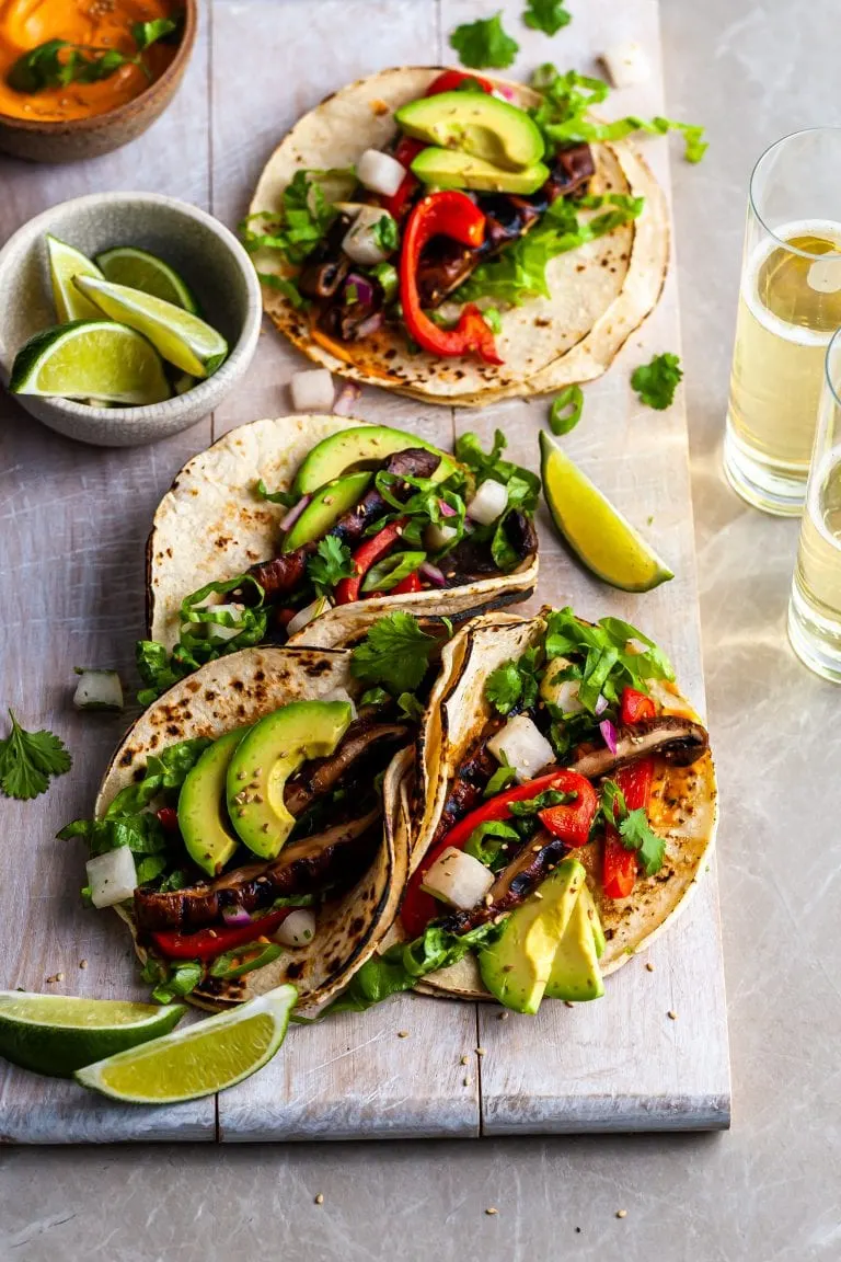4 Korean Mushroom Tacos on a white serving board served with lime wedges, Gochujang crema and beer
