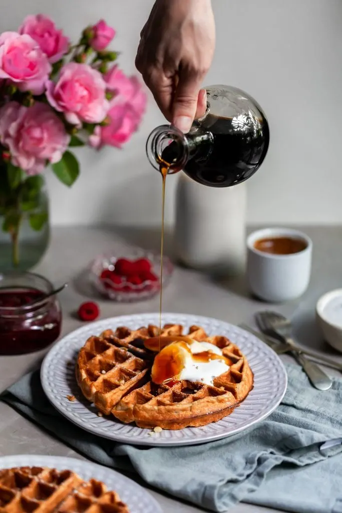 Vegan Banana Waffles served with vegan cream, fresh bananas and a bottle of maple syrup being poured over them.