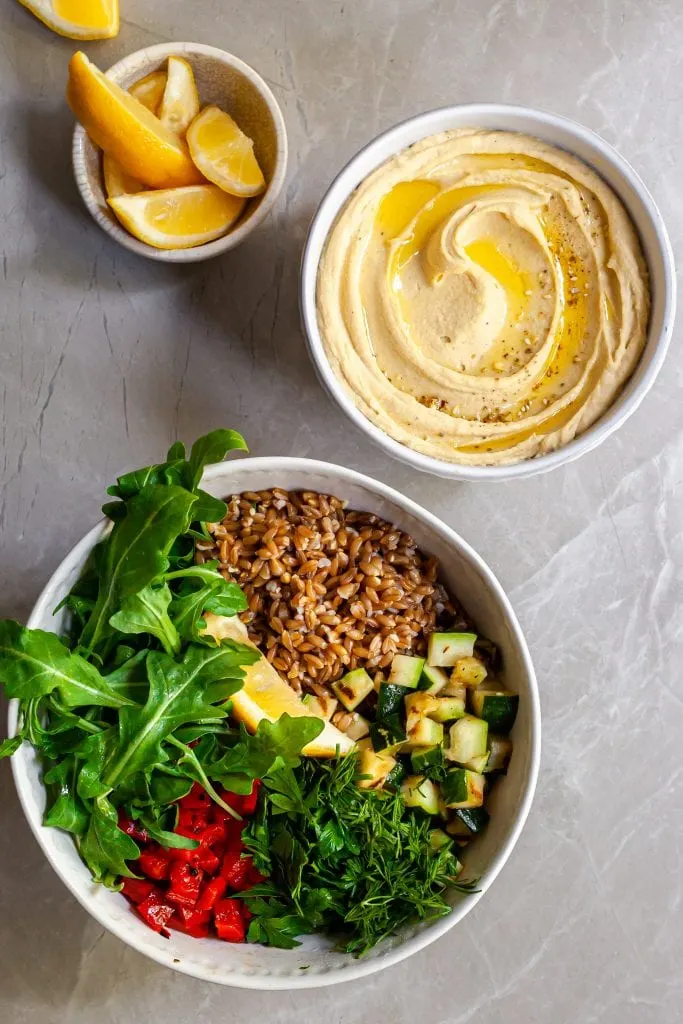 Ingredients for the farro salad in a large bowl before tossing, hummus in  a second bowl and lemon wedges in a third bowl.