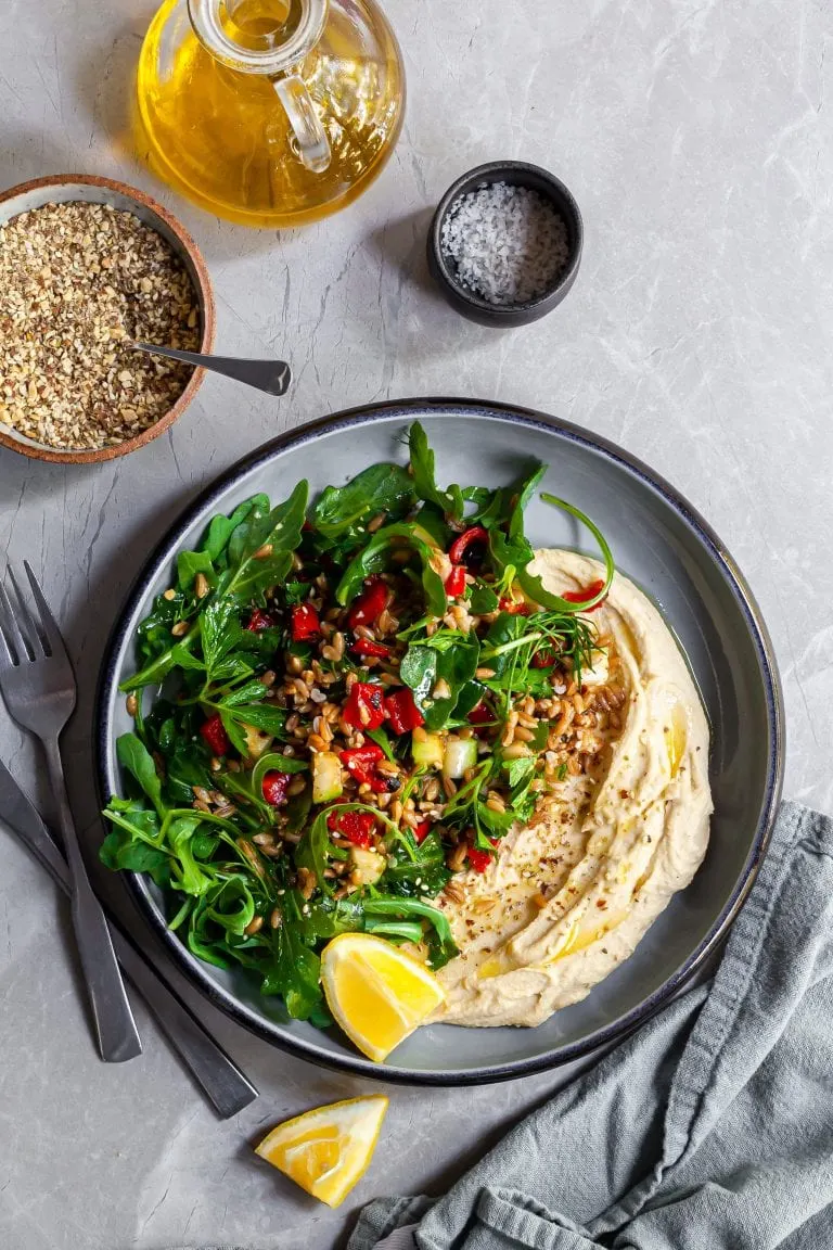 Looking down on a bowl of farro salad over hummus, served with lemon wedges and dukkah