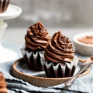 Two chocolate cupcakes on a plate with a bowl of fluffy chocolate ganache frosting on the side
