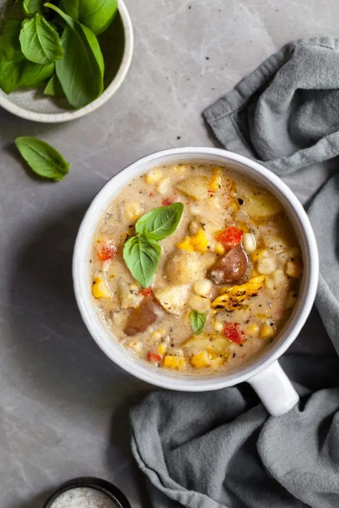 Flatlay of a large white mug filled with vegan corn and potato chowder garnished with fresh basil
