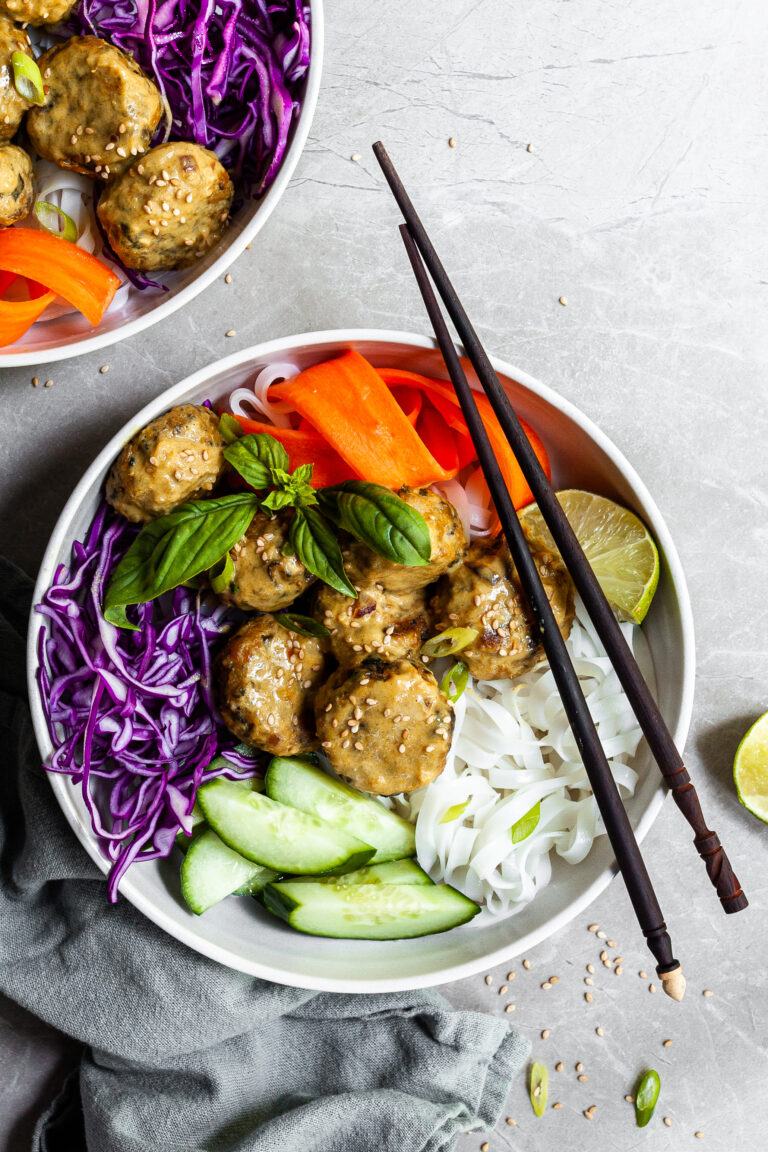 White bowl and chopsticks with rice noodles, chcikpea balls covered in yellow curry sauce, and raw veggies