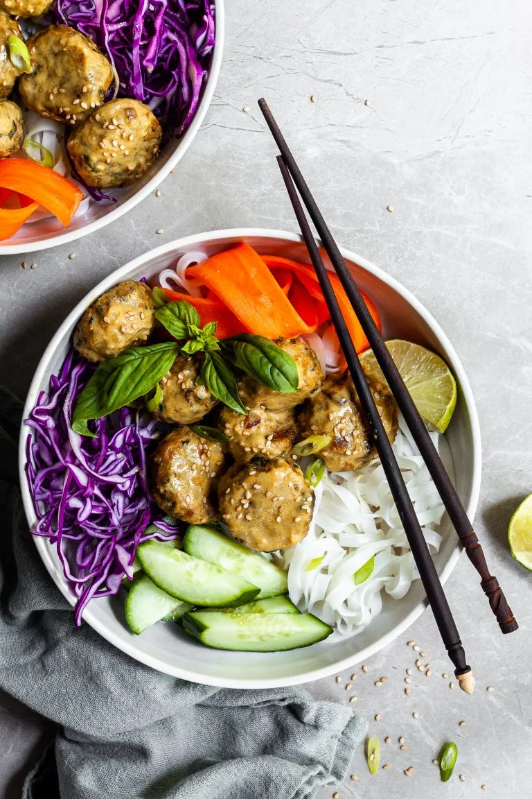 White bowl and chopsticks with rice noodles, chcikpea balls covered in yellow curry sauce, and raw veggies