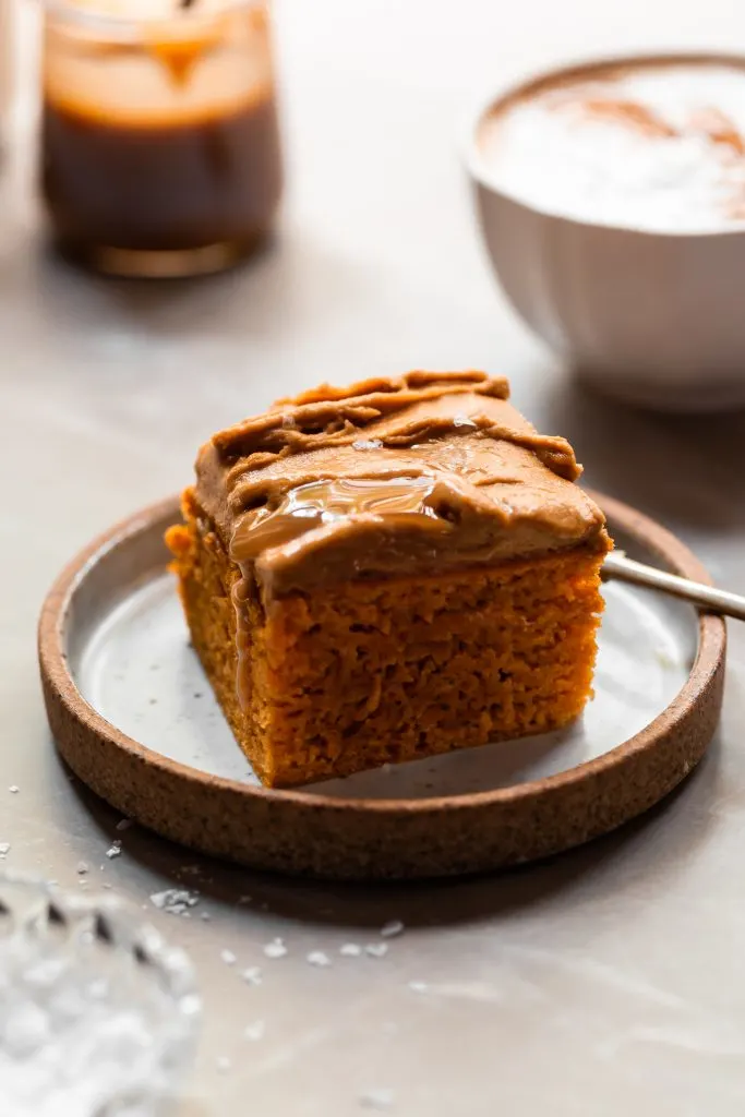 A slice of pumpkin spice sheetcake topped with epsresso buttercream and a drizzle of butterscotch sauce, served with a latte and a jar of butterscotch in background