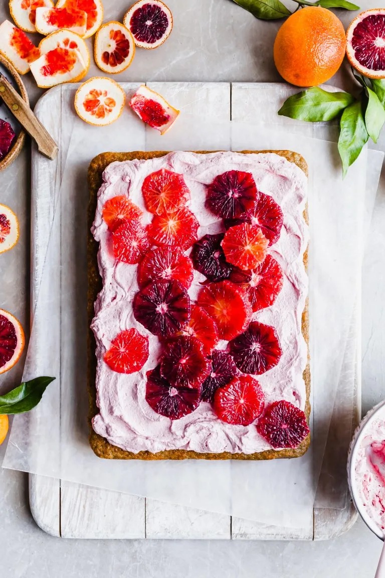 Flatlay of a sheet cake covered in pink frosting and slices of blood oranges, the background littered with orange rinds