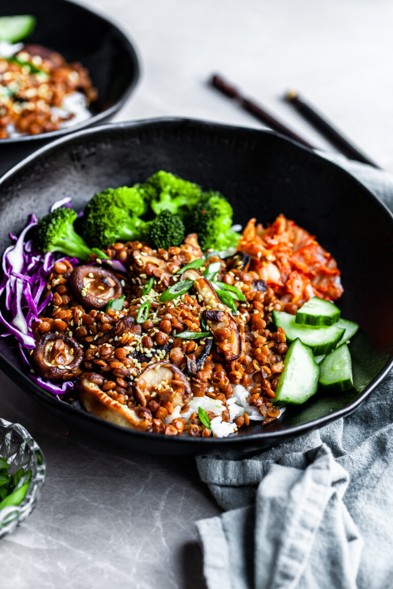 Angled view of a black bowl filled with rice topped with lentils, shiitakes, broccoli and kimchi