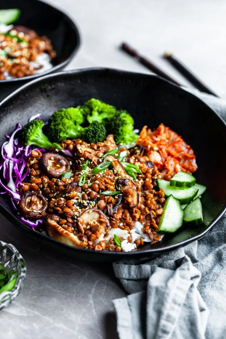 Angled view of a black bowl filled with rice topped with lentils, shiitakes, broccoli and kimchi