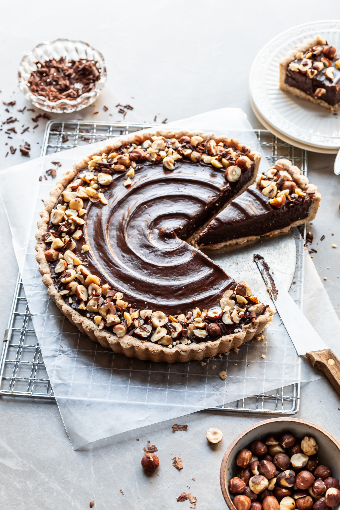 Angled view of a chocolate tart  on a cooling rack garnished with hazelnuts. Slices have been served and one slice sits waiting to be plated. 