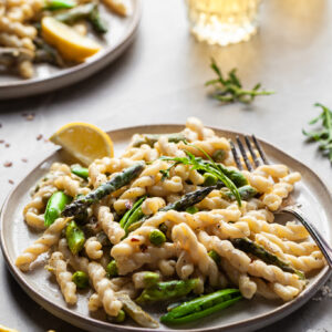 Side view of a plate of gemelli pasta in a light cream sauce with asparagus and spring peas with glasses of wine in the background