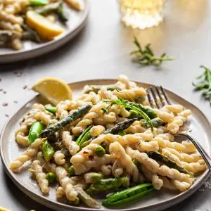 Side view of a plate of gemelli pasta in a light cream sauce with asparagus and spring peas with glasses of wine in the background