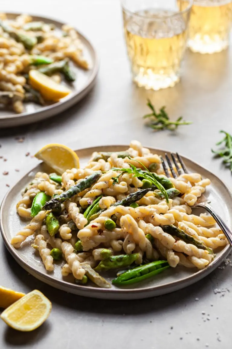 Side view of a plate of gemelli pasta in a light cream sauce with asparagus and spring peas with glasses of wine in the background