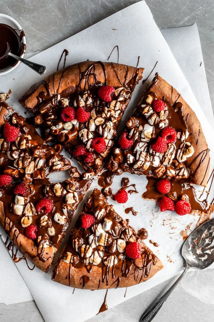 vegan rocky road dessert pizza garnished with raspberries is cut into slices, drizzled with chocolate sauce and placed on a piece of white parchment paper