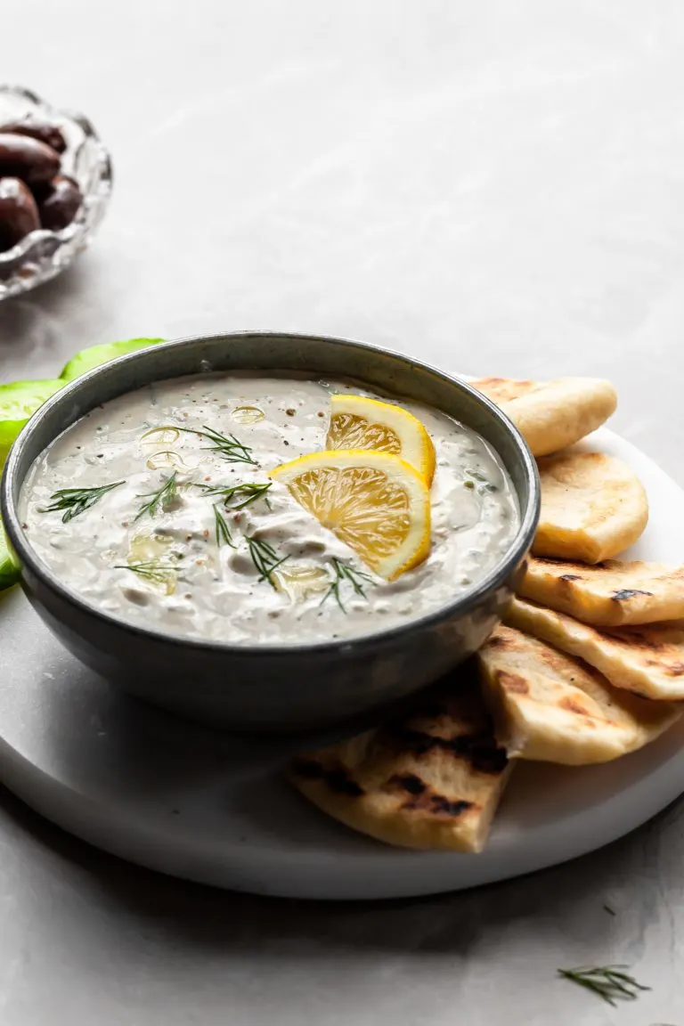 Grey bowl of tzatziki sauce garnished with lemon wedges, served with greek flatbread and slices of cucumber