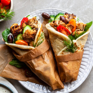 2 cauliflower gyro wraps wrapped in brown paper on a plate beside a bowl of tzatziki