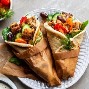 2 cauliflower gyro wraps wrapped in brown paper on a plate beside a bowl of tzatziki