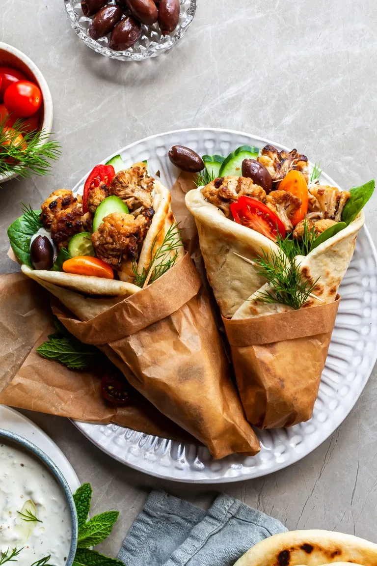 2 cauliflower gyro wraps wrapped in brown paper on a plate beside a bowl of tzatziki