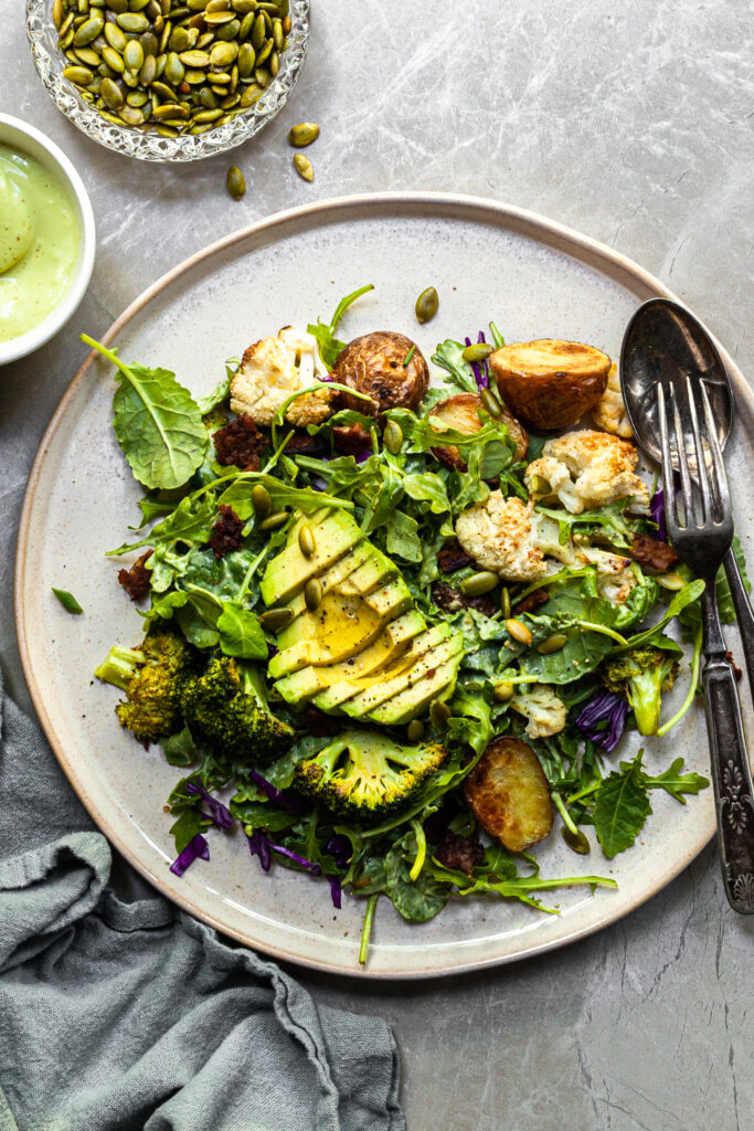 A serving of kale and roasted vegetable salad on an aluminium tray served with a small white bowl filled with creamy light green avocado Caesar dressing