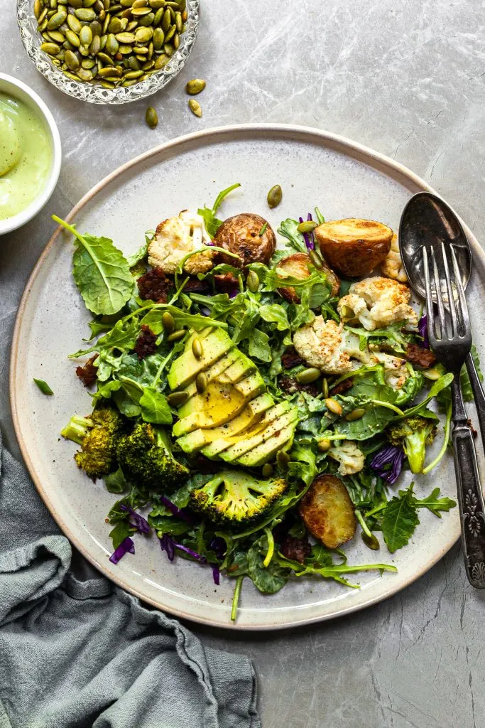 A serving of kale and roasted vegetable salad on an aluminium tray served with a small white bowl filled with creamy light green avocado Caesar dressing