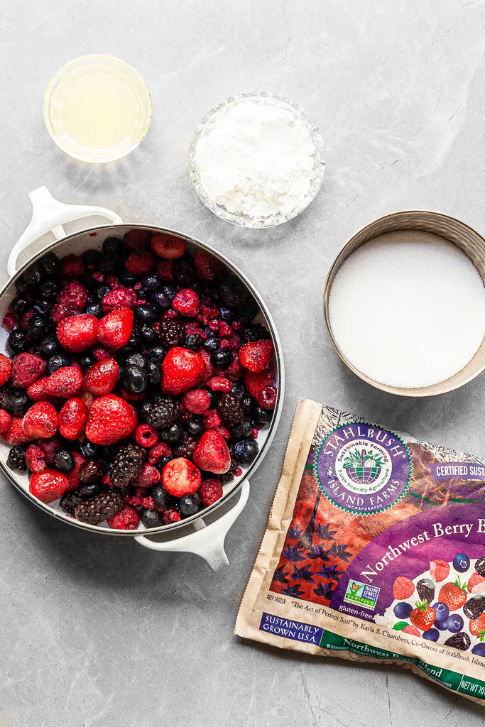 A white pot filled with a mixture of frozen berries, a bowl of sugar, cornstarch and lemon juice beside a bag of Stahlbush Island Farms Northwest Berry Blend