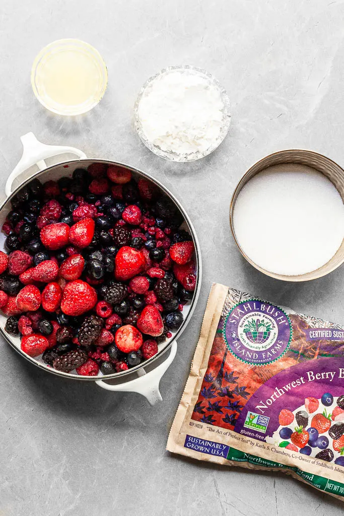 A white pot filled with a mixture of frozen berries, a bowl of sugar, cornstarch and lemon juice beside a bag of Stahlbush Island Farms Northwest Berry Blend