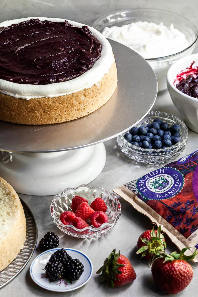 The first layer of cake on a cake stand, covered in a layer of vanilla frosting and berry curd held in with a frosting dam. Bowls of fresh fruit sit beside the cake being assembled. 