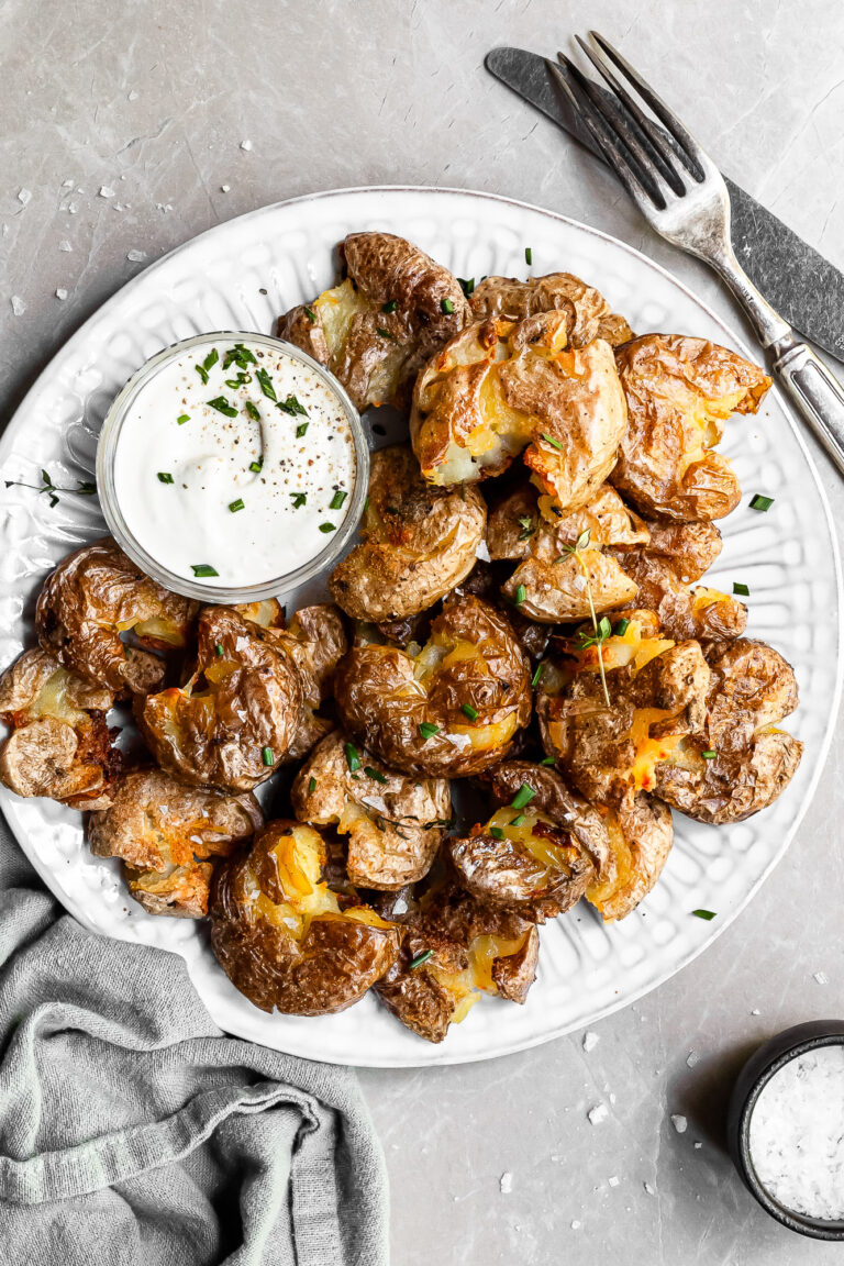White plate filled with crispy smashed potatoes and a small glass bowl of vegan sour cream