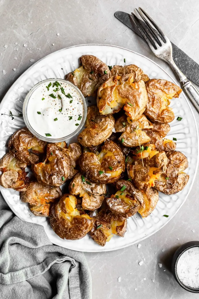 White plate filled with crispy smashed potatoes and a small glass bowl of vegan sour cream