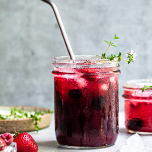 Jar filled with vibrant purplish reddish fizzy drink, garnished with thyme sprigs and served with a straw.