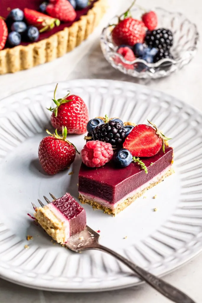 A bite of berry chocolate tart rests on a fork with the rest of the purple berry tart in the background on a white plate