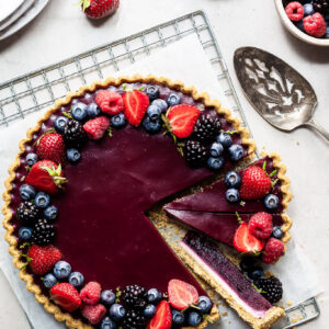 Flaylay of a purple tart with shortbread crust covered in summer berries. 3 slices are made with one on its side revealing the white chocolate ganache on the bottom.