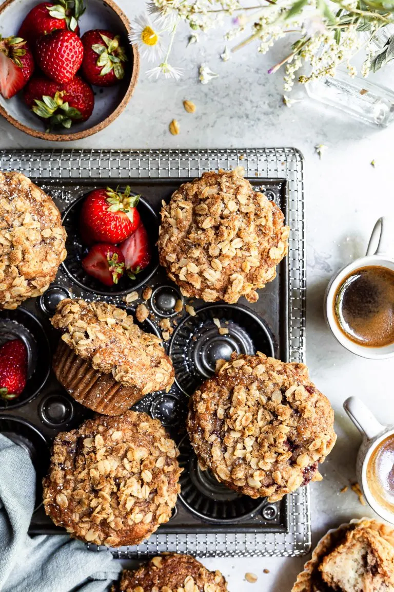 View looking down on a baking tray of muffins beside a bowl of strawberries, a vase of wildflowers and 2 some cups of espresso