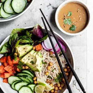 white bowl filled with rice noodles, cubes of sweet potatoes, chickpeas and cucumbers, with a small bowl of tahini dressing on the side